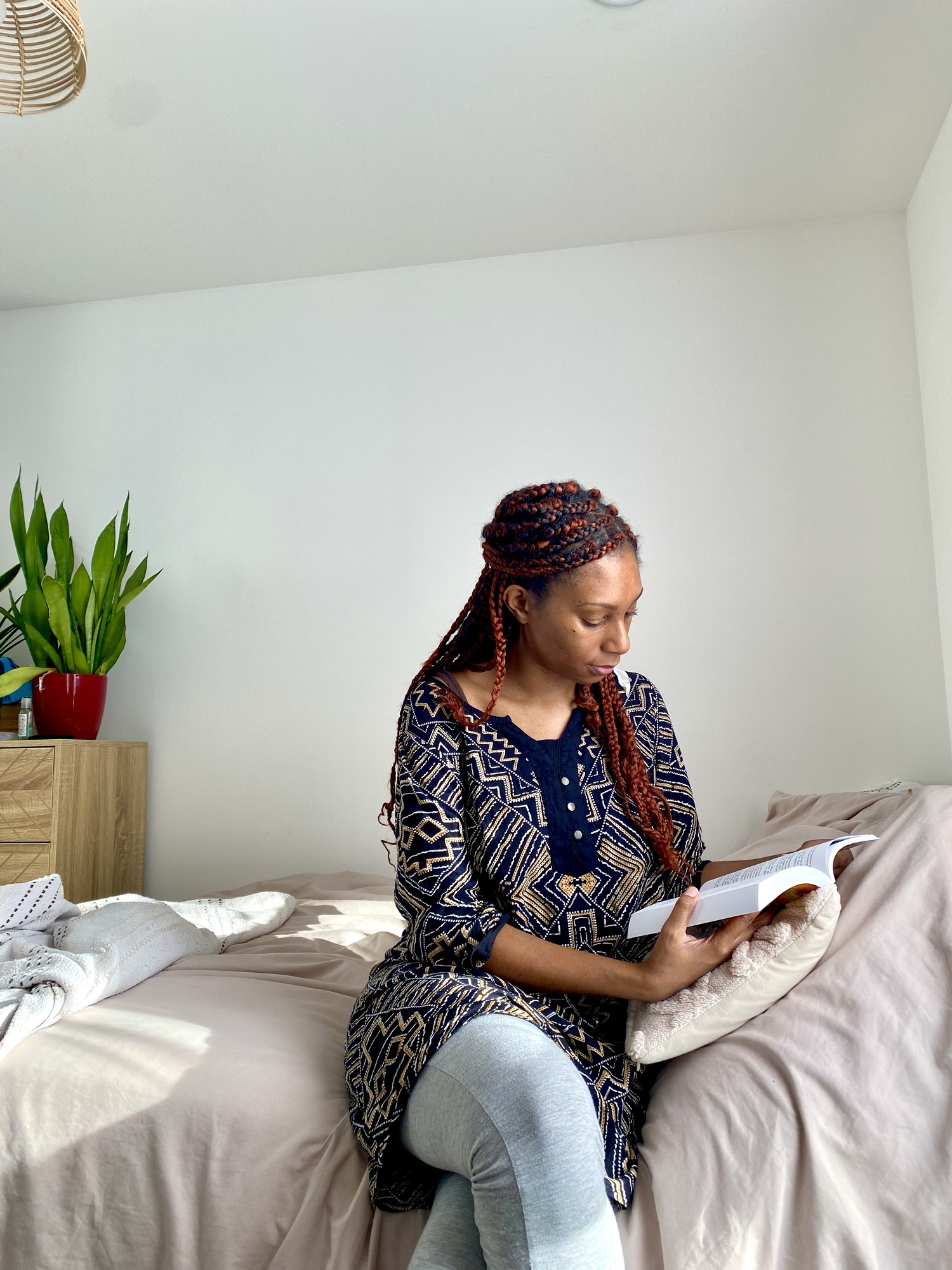 A Black woman sits on a bed reading a book.